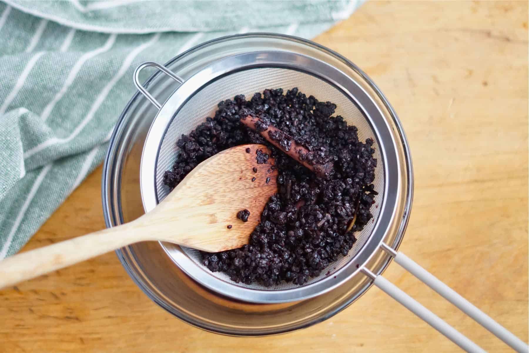 wooden spoon in sieve with cooked elderberries on top of bowl