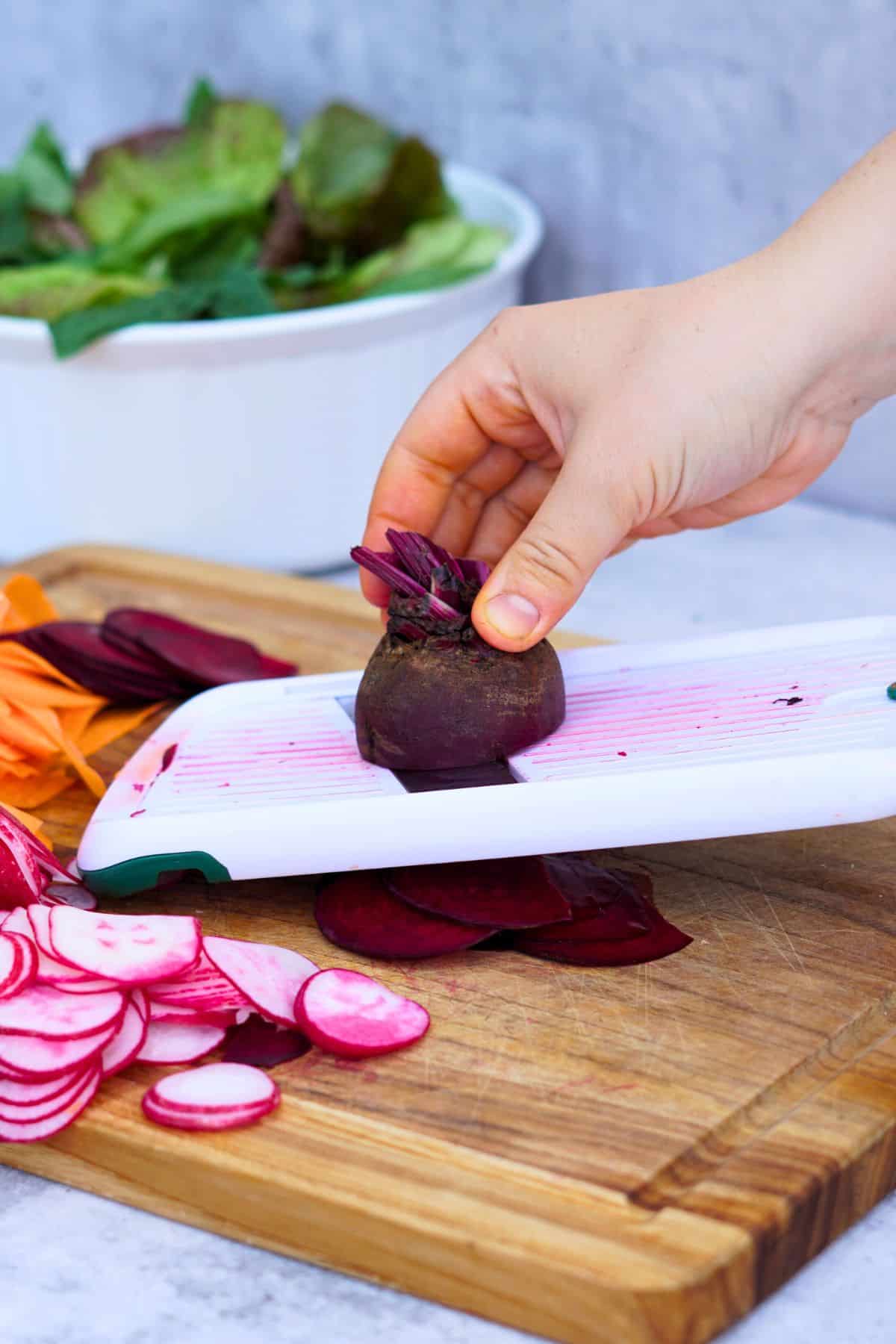 hand slicing beets on a mandolin next to sliced carrots and radish