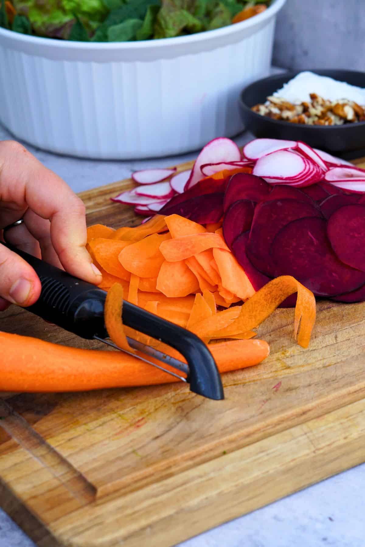 hand peeling carrots on cutting board next to sliced beets and radish