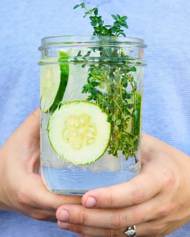hands holding glass jar of ice water with sliced cucumber and thyme sprigs