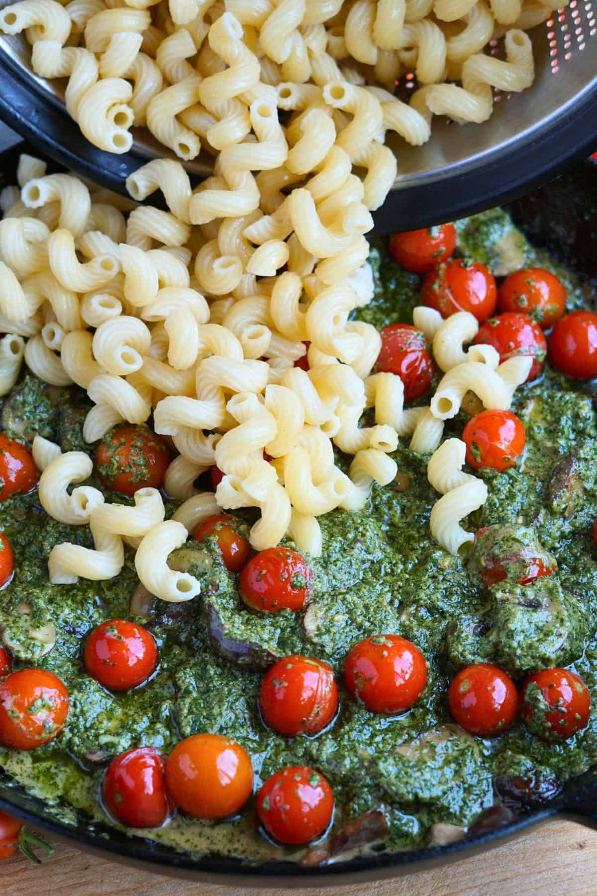 cavatappi pasta pouring into cast iron pan with pesto, mushrooms, and tomatoes