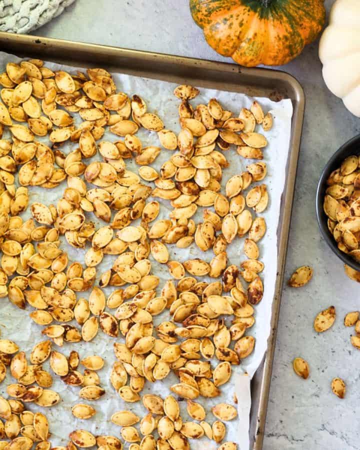 roasted pumpkin seeds on a baking sheet next to small serving dish and small pumpkins