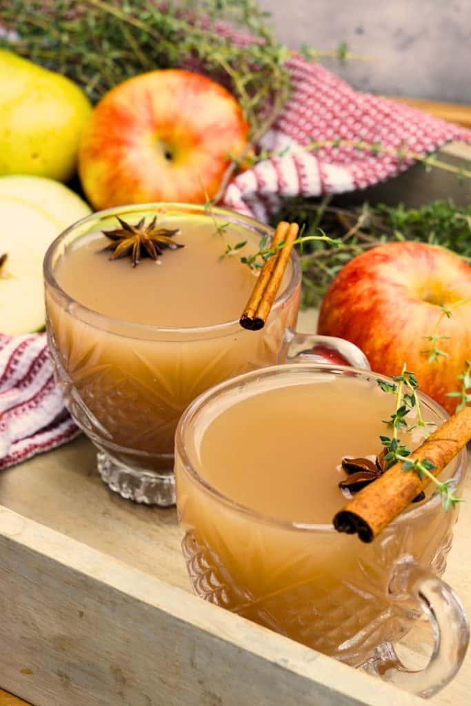 two glasses of spiced apple scrap cider garnished with cinnamon sticks and star anise on tray with whole apples