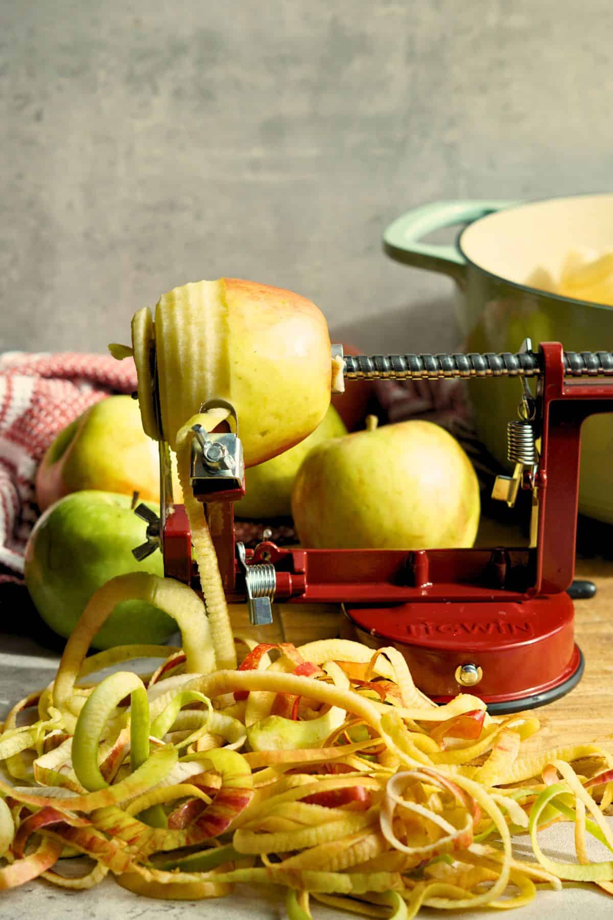 An apple being peeled, sliced, and cored with pot and whole apples in the background
