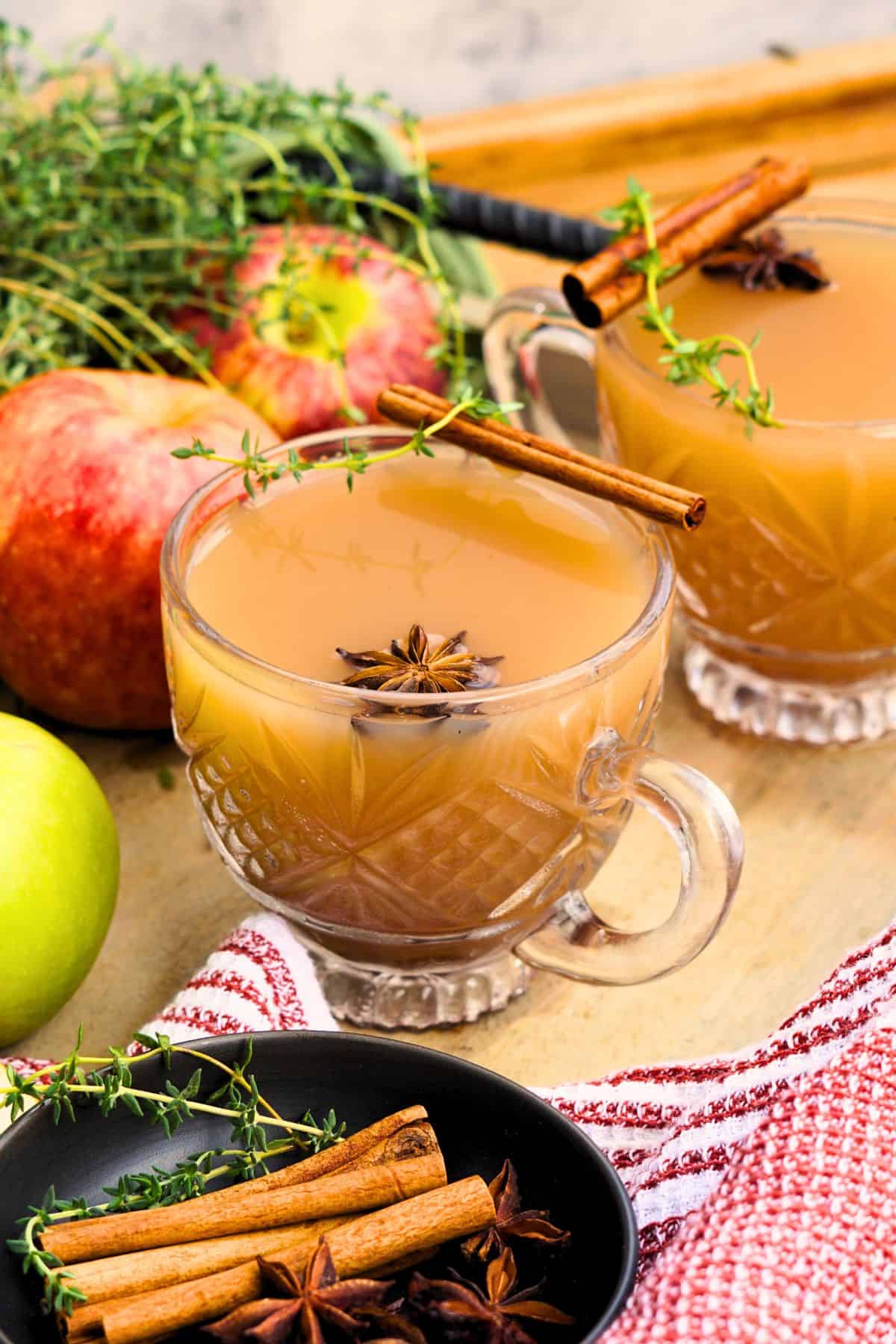two glasses of spiced apple scrap cider garnished with cinnamon sticks and star anise on tray with whole apples
