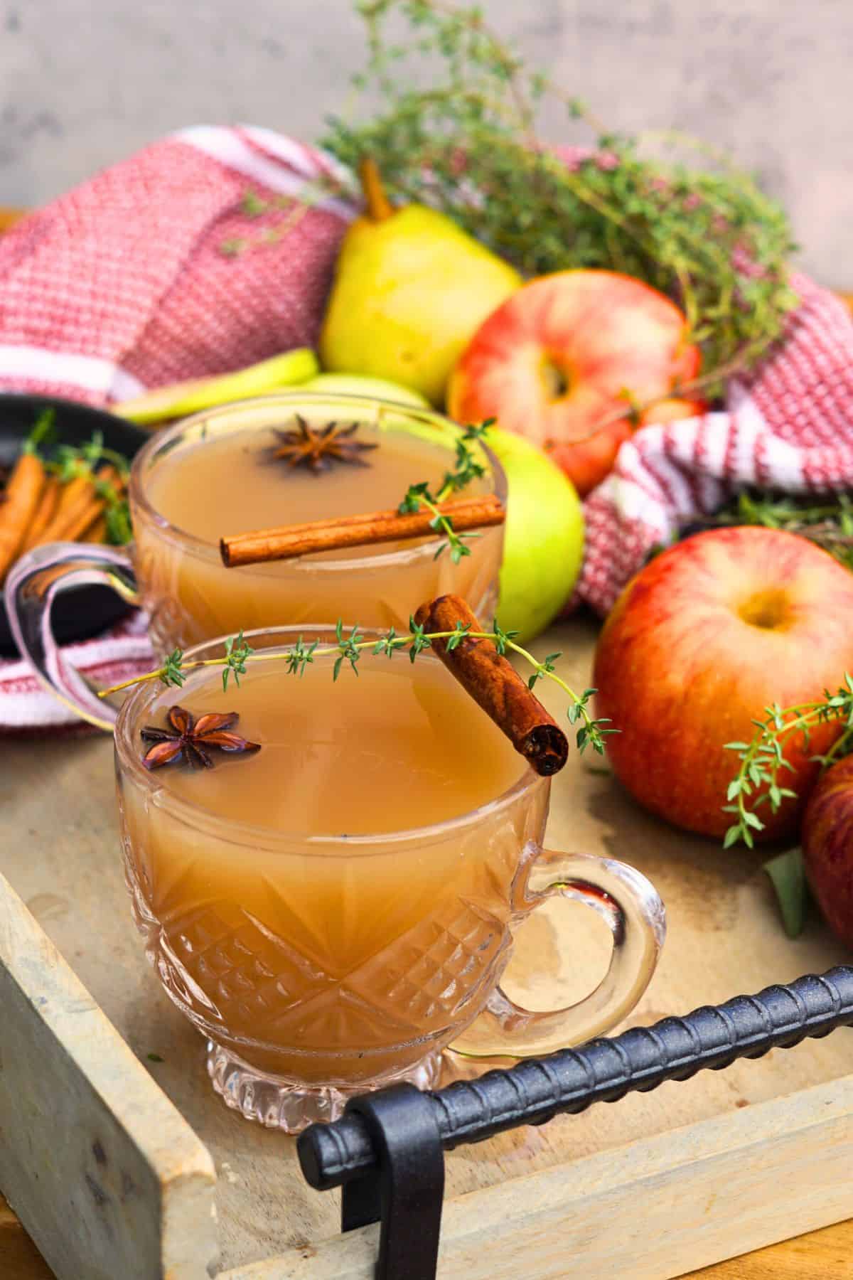 two glasses of spiced apple scrap cider garnished with cinnamon sticks and star anise on tray with whole apples
