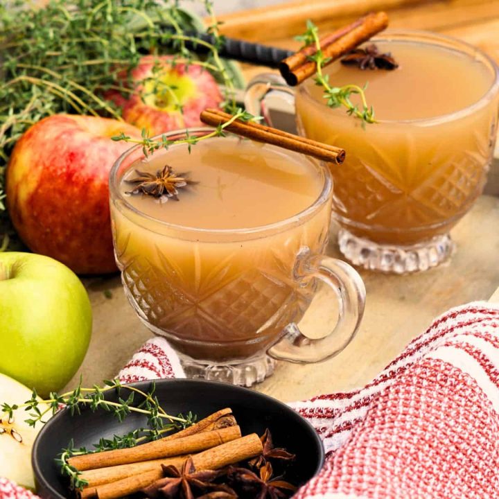 two glasses of spiced apple scrap cider garnished with cinnamon sticks and star anise on tray with whole apples