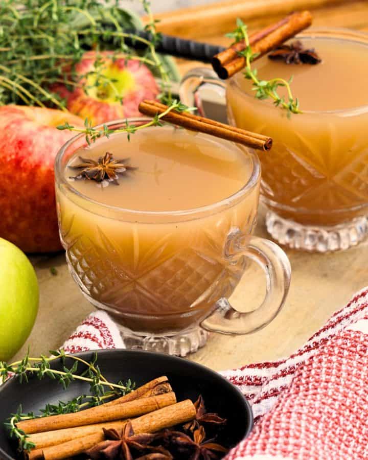 two glasses of spiced apple scrap cider garnished with cinnamon sticks and star anise on tray with whole apples