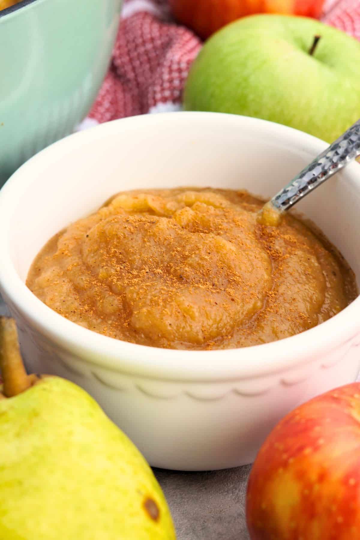 spiced pear applesauce in a small white bowl with a spoon in counter next to whole apples and pears